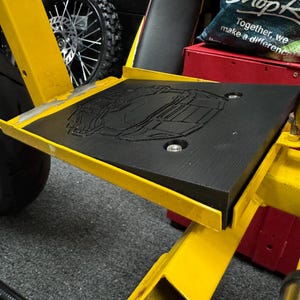 Puede incluir: Un marco de metal amarillo con un reposapiés de goma negra. El reposapiés tiene un diseño en relieve de un camión. El marco tiene una manguera negra unida a él.