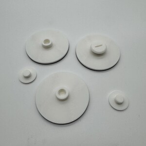 Peut inclure: Plusieurs objets circulaires blancs avec un liser&eacute; noir. Les objets varient en taille et pr&eacute;sentent une partie centrale sur&eacute;lev&eacute;e. Certains ont un trou, d'autres une fente. Ils sont dispos&eacute;s sur une surface blanche.