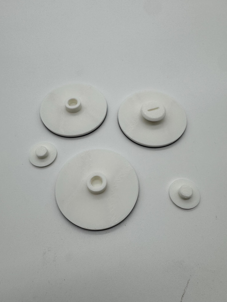 Peut inclure: Plusieurs objets circulaires blancs avec un liser&eacute; noir. Les objets varient en taille et pr&eacute;sentent une partie centrale sur&eacute;lev&eacute;e. Certains ont un trou, d'autres une fente. Ils sont dispos&eacute;s sur une surface blanche.