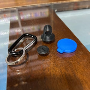 Pu&ograve; includere: Un moschettone nero con un anello argentato, un bottone rotondo nero, un bottone rotondo blu e un oggetto 3D stampato in nero a forma di figura incappucciata.