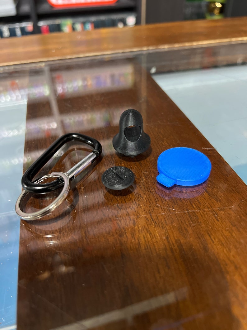 Pu&ograve; includere: Un moschettone nero con un anello argentato, un bottone rotondo nero, un bottone rotondo blu e un oggetto 3D stampato in nero a forma di figura incappucciata.