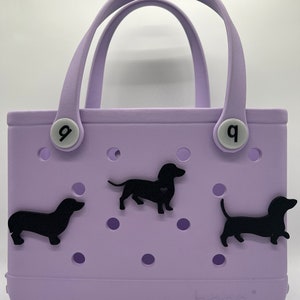 May include: A lavender-colored tote bag with black dachshund silhouettes and the word "BOGG" printed on the bottom. The bag has two handles and two white buttons with the number "9" on them.
