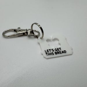Può includere: Portachiavi in plastica bianca con un anello e un moschettone in metallo argentato. Il portachiavi ha la forma di una pagnotta di pane ritagliata e il testo "Let's get this bread".