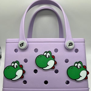 Puede incluir: Un bolso tote lila con tres dise&ntilde;os de dinosaurios verdes de dibujos animados. El bolso tiene un asa y la marca "bogg" es visible en la parte delantera.
