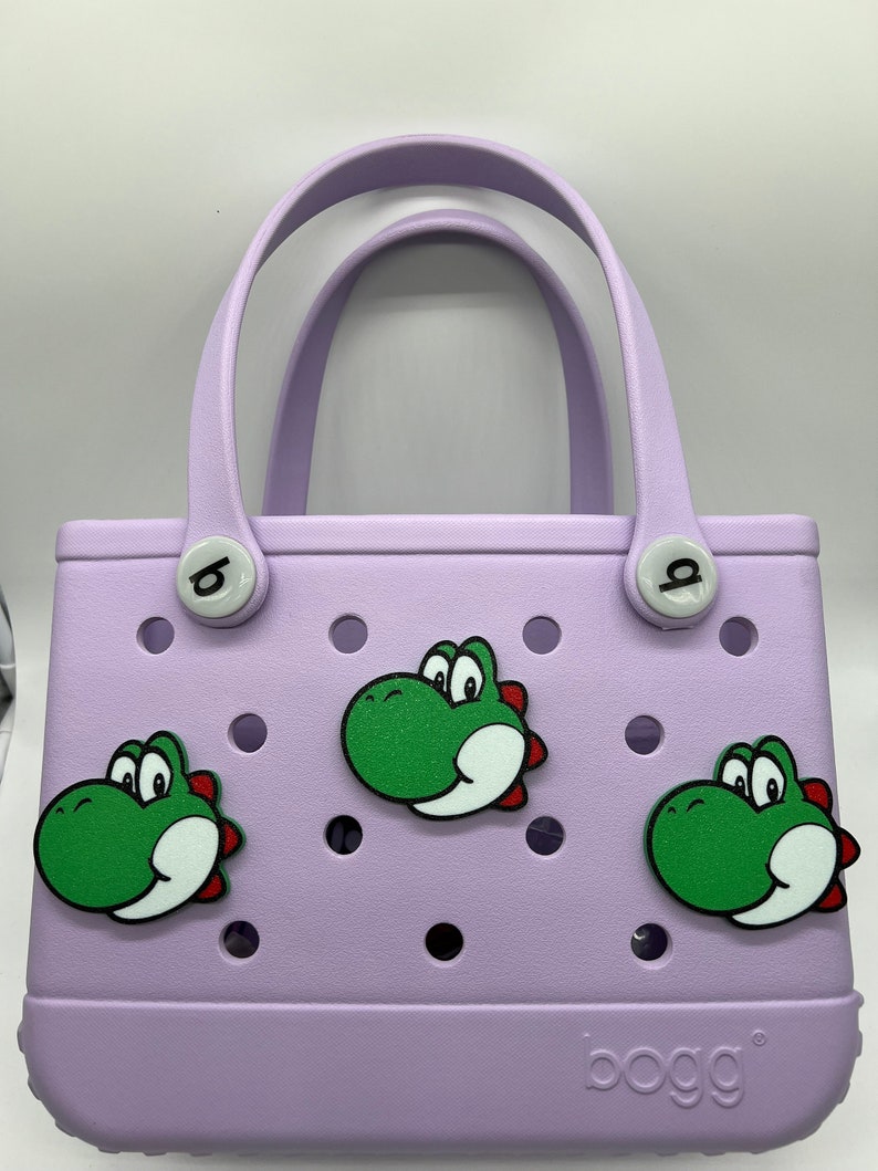 Puede incluir: Un bolso tote lila con tres dise&ntilde;os de dinosaurios verdes de dibujos animados. El bolso tiene un asa y la marca "bogg" es visible en la parte delantera.