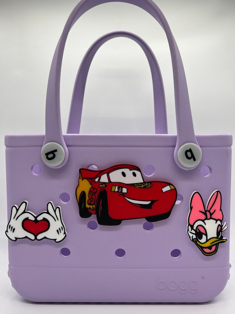 Peut inclure: Un sac fourre-tout lavande avec une voiture rouge de Flash McQueen, un c&oelig;ur blanc avec des mains et un personnage de Daisy Duck rose et jaune. Le sac a un logo blanc qui dit "oooh!" en bas.