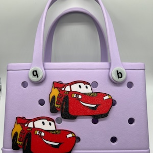 Peut inclure: Un sac Bogg lavande avec deux breloques de voiture rouge &Eacute;clair McQueen. Le sac a deux poign&eacute;es blanches et le mot "bogg" est imprim&eacute; au bas.