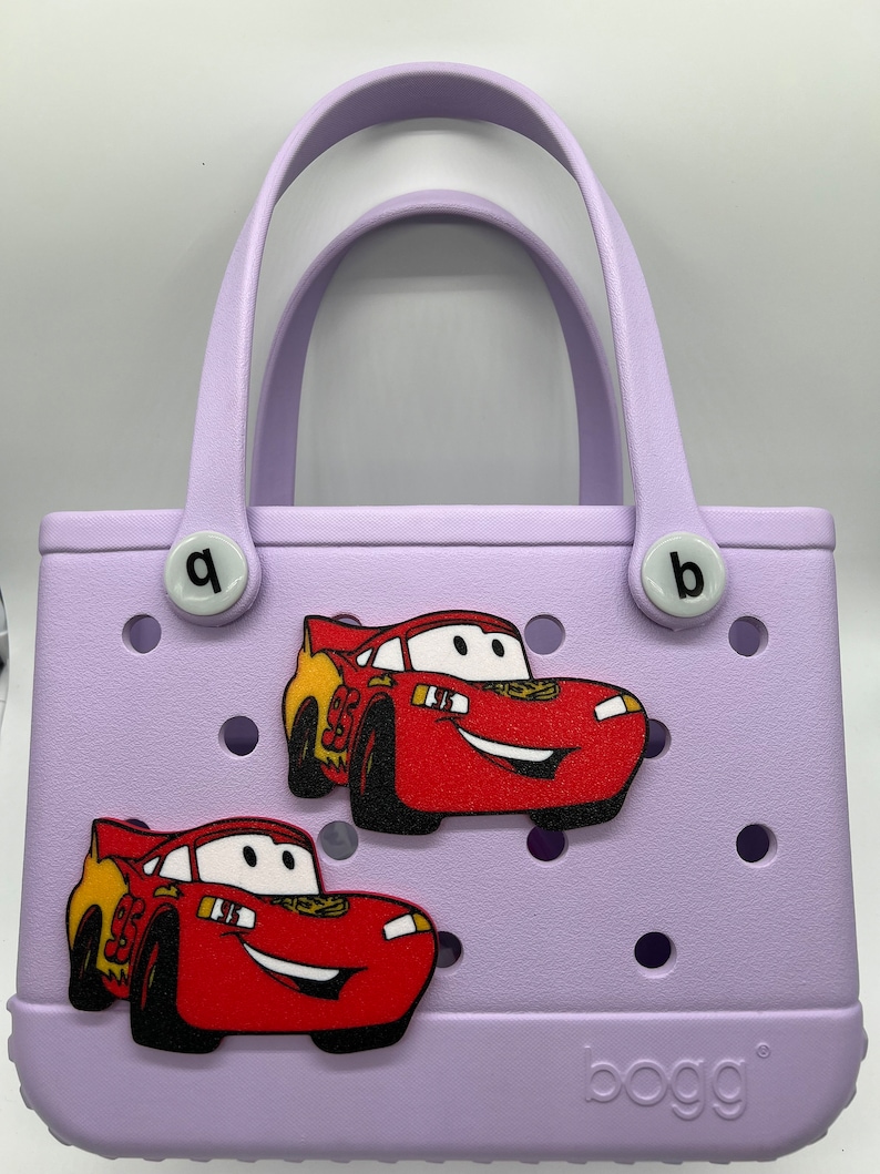 Peut inclure: Un sac Bogg lavande avec deux breloques de voiture rouge &Eacute;clair McQueen. Le sac a deux poign&eacute;es blanches et le mot "bogg" est imprim&eacute; au bas.