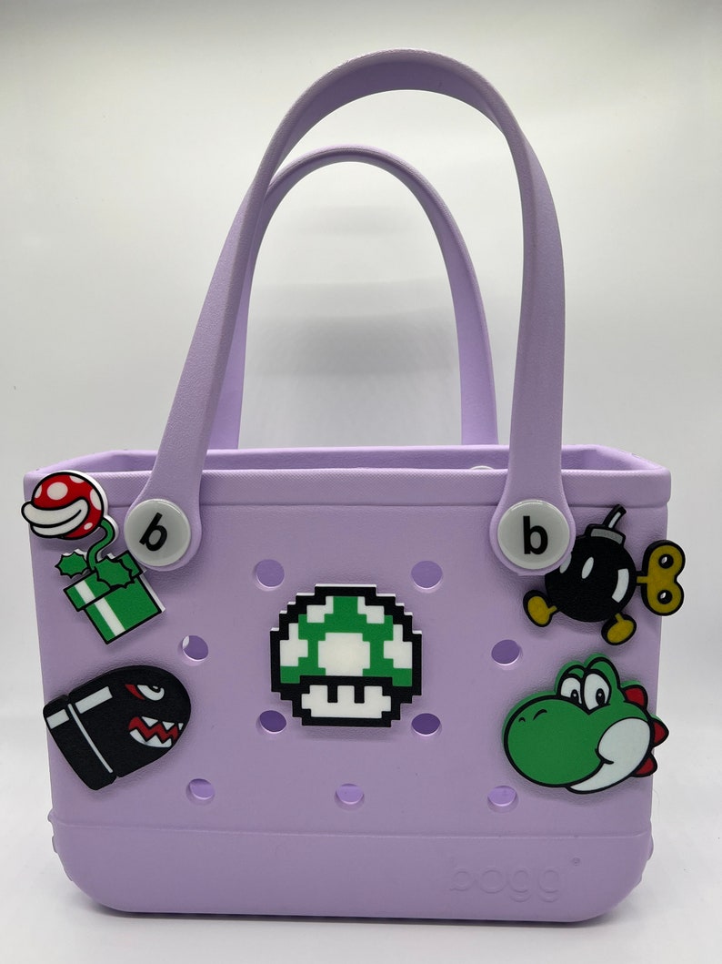 Puede incluir: Un bolso tote lavanda con dos asas y varios amuletos de personajes de dibujos animados. Los amuletos incluyen un hongo verde, una bala negra, un Yoshi verde y una bomba negra. La bolsa tiene la palabra "bogg" impresa en la parte inferior.