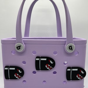 Puede incluir: Un bolso tote lila con dos asas y tres abalorios negros con forma de bala con detalles rojos y blancos. El bolso tiene dos botones circulares blancos con las letras "b" y "q". La marca "bogg" est&aacute; impresa en la parte inferior.