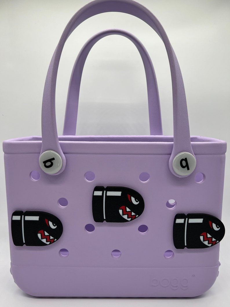 Puede incluir: Un bolso tote lila con dos asas y tres abalorios negros con forma de bala con detalles rojos y blancos. El bolso tiene dos botones circulares blancos con las letras "b" y "q". La marca "bogg" est&aacute; impresa en la parte inferior.