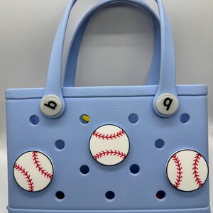 Peut inclure: Un sac fourre-tout bleu clair avec deux poign&eacute;es et des d&eacute;corations sur le th&egrave;me du baseball. Le sac pr&eacute;sente trois motifs de balles de baseball avec des coutures rouges et les lettres "b" et "q" sur les attaches de la poign&eacute;e. La marque "bogg" est visible.