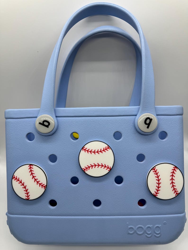 Peut inclure: Un sac fourre-tout bleu clair avec deux poign&eacute;es et des d&eacute;corations sur le th&egrave;me du baseball. Le sac pr&eacute;sente trois motifs de balles de baseball avec des coutures rouges et les lettres "b" et "q" sur les attaches de la poign&eacute;e. La marque "bogg" est visible.