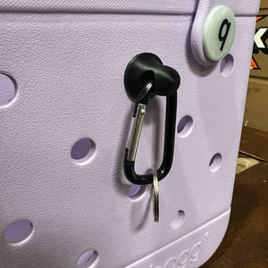 Pu&ograve; includere: Una borsa tote di colore lavanda con un moschettone nero attaccato a un gancio nero. La borsa ha un cerchio bianco con il numero "9" su di esso. La borsa ha la parola "BOGG" stampata sul fondo.