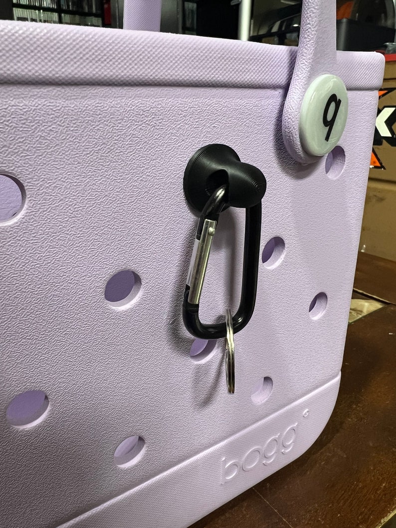 Pu&ograve; includere: Una borsa tote di colore lavanda con un moschettone nero attaccato a un gancio nero. La borsa ha un cerchio bianco con il numero "9" su di esso. La borsa ha la parola "BOGG" stampata sul fondo.