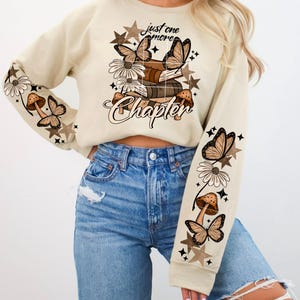 May include: Beige sweatshirt with a graphic design featuring butterflies, mushrooms, daisies, and stars. The text "just one more Chapter" is printed on the front. The sleeves have repeating patterns of mushrooms, butterflies, and daisies.