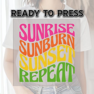 May include: A white t-shirt with a graphic design that says "Sunrise Sunburn Sunset Repeat" in a retro font with colors that transition from pink to orange to yellow to green.