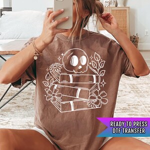 May include: A brown t-shirt with a white design of a skull sitting on top of a stack of books with flowers around the design. The text "READY TO PRESS DTF TRANSFER" is at the bottom of the image.