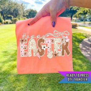 May include: Coral-colored DTF transfer with the words "Happy Easter" in a decorative font. The letters are adorned with floral and Easter-themed graphics, including a carrot and an egg. The transfer is ready to press.