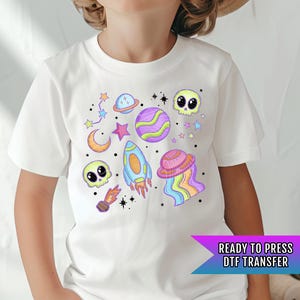 May include: White t-shirt with a colorful space-themed design. The design includes planets, stars, a rocket, a skull, and a crescent moon. The text "READY TO PRESS DTF TRANSFER" is visible in the bottom right corner.