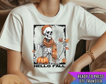 Pode incluir: Camiseta branca com um gráfico de um esqueleto usando um gorro e uma echarpe segurando uma abóbora e folhas. O texto "Hello Fall" está abaixo do gráfico.