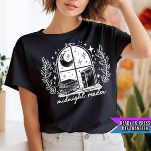 May include: Black t-shirt with a white graphic design of a window with a crescent moon, stars, and books. The text "midnight reader" is below the window.