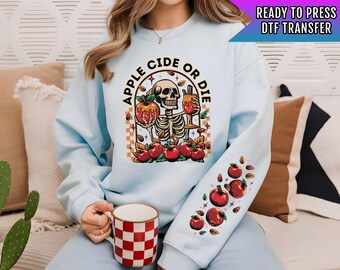 May include: Light blue sweatshirt with a graphic of a skeleton holding an apple cider drink and the text "Apple Cider or Die". The sweatshirt has a checkered pattern and red apples on the sleeves.