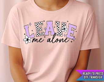 May include: Pink t-shirt with a checkered design and the text "LEAVE me alone" in purple with small flower accents.