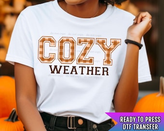 May include: White t-shirt with a brown and peach plaid design that says "COZY WEATHER" in a blocky font.