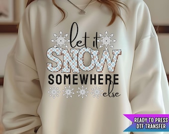 May include: A cream colored sweatshirt with the text "Let it snow somewhere else" in black. The text is surrounded by snowflake designs in blue and brown. The sweatshirt is a ready to press DTF transfer.