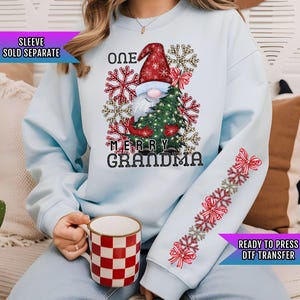 May include: Light blue sweatshirt featuring a Christmas gnome holding a tree, with the text "ONE MERRY GRANDMA." The sleeve has a snowflake and bow design. A red and white checkered mug is held in the hand.