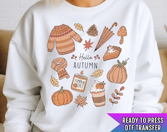 Pode incluir: Camisola branca com um design gráfico de temática outonal. O design inclui ilustrações de um suéter, uma echarpe, uma vela de especiarias de abóbora, uma abóbora, folhas, uma torta, um cogumelo, uma pinha e um guarda-chuva. O texto "Hello Autumn" está incluído no design.