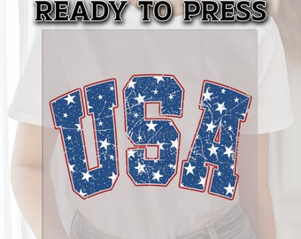 May include: A white t-shirt with a distressed blue and red "USA" graphic featuring white stars. The graphic is ready to be pressed onto a shirt.