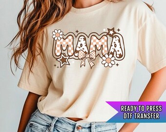May include: Cream-colored t-shirt with the word "MAMA" in a retro font, decorated with flowers, stars, and a bow. The letters have a checkered pattern. A purple and white banner at the bottom reads "READY TO PRESS DTF TRANSFER".