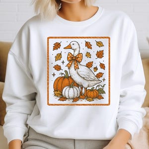 May include: White sweatshirt featuring a fall-themed graphic. The design includes a white goose with an orange bow, pumpkins, and autumn leaves. The graphic is framed with an orange border, creating a seasonal and festive look.