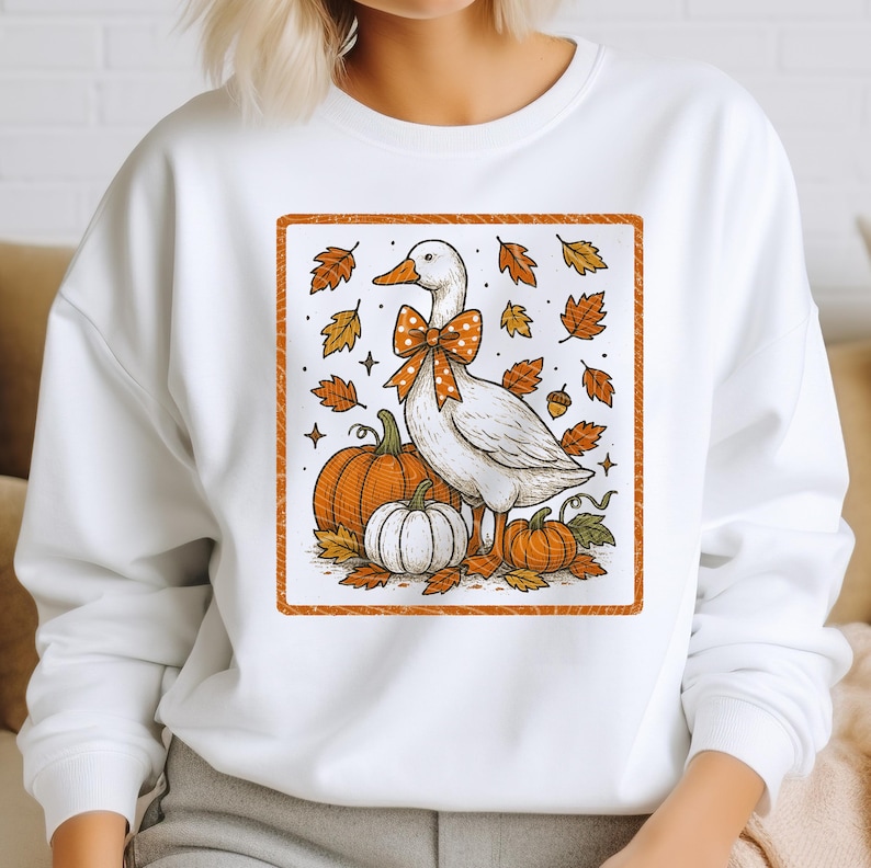 May include: White sweatshirt featuring a fall-themed graphic. The design includes a white goose with an orange bow, pumpkins, and autumn leaves. The graphic is framed with an orange border, creating a seasonal and festive look.