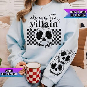 May include: Light blue sweatshirt with a black and white checkered pattern and the text "always the villain" with a dripping apple design. The apple has a skull face. The sleeve has a smaller version of the apple design.