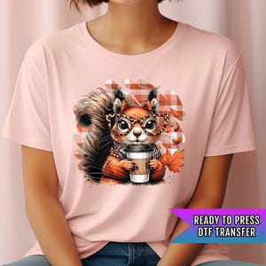 May include: A light pink t-shirt featuring a cartoon squirrel wearing glasses and holding a coffee cup. The squirrel is set against a plaid background with fall leaves. The text "READY TO PRESS DTF TRANSFER" is in a purple and blue banner.