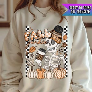 May include: A white sweatshirt with a graphic design of a skeleton holding a cup of coffee and pumpkins. The text "Fall Junkie" is written in black letters above the skeleton. The design is surrounded by a checkered pattern.