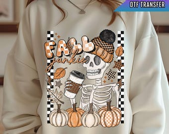 Pode incluir: Uma camisa de moletom branca com um design gráfico de um esqueleto segurando uma xícara de café e abóboras. O texto "Fall Junkie" está escrito em letras pretas acima do esqueleto. O design é cercado por um padrão xadrez.