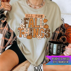 May include: A beige t-shirt with the text "ALL THE FALL THINGS" in orange and brown, featuring pumpkins, flowers, and leaves. The shirt is paired with a black mug with white ghost designs. A "READY TO PRESS DTF TRANSFER" banner is visible.