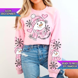 May include: Pink sweatshirt with a snowman graphic and the text "Baby, it's cold outside". The snowman is wearing a pink hat and scarf. The sweatshirt has snowflake designs on the sleeves.