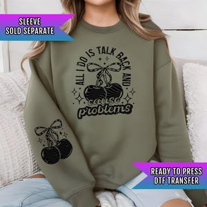 May include: Olive green sweatshirt with a black cherry graphic and the text "ALL I DO IS TALK BACK AND CAUSE PROBLEMS." The sleeve has a cherry graphic. The text "SLEEVE SOLD SEPARATE" and "READY TO PRESS DTF TRANSFER" are also visible.