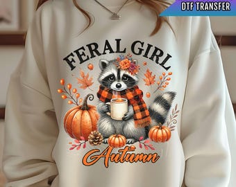 May include: A white sweatshirt with a graphic design of a raccoon wearing a plaid scarf and holding a cup of coffee. The design also includes pumpkins, fall leaves, and the text "Feral Girl Autumn".