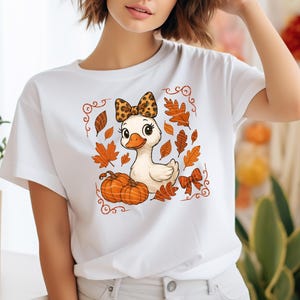 May include: White t-shirt featuring a cartoon duck with a leopard print bow, surrounded by fall leaves and pumpkins. The design has a decorative border. The duck has large eyes and a cute expression. The shirt is a casual style.