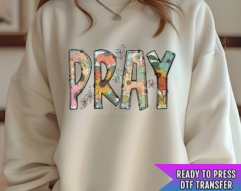 May include: A cream-colored sweatshirt with a floral design that spells out the word "PRAY". The sweatshirt is a ready-to-press DTF transfer.