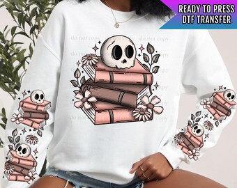 Könnte beinhalten: Ein weißer Pullover mit einem rosa und braunen Stapel Bücher, die mit einem Schädel und Blumen verziert sind. Die Ärmel haben ein sich wiederholendes Muster des gleichen Designs.