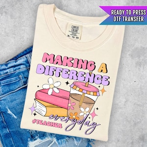 May include: A beige t-shirt with a pink and purple graphic that says "Making a Difference Everyday #Teacher". The graphic also includes a stack of books, a cup of coffee, and flowers.