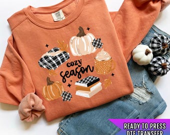 May include: An orange sweatshirt with a fall-themed graphic that includes pumpkins, a coffee cup, and the words "cozy season." The sweatshirt is paired with blue jeans and decorative pumpkins. The graphic is a DTF transfer.