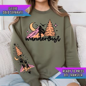 May include: A green sweatshirt with a graphic design of a mountain range, trees, a crescent moon, and stars. The text "wanderlust" is printed in black below the graphic. The sweatshirt has a long sleeve with a graphic design of an arrow, a pine tree, and paw prints.